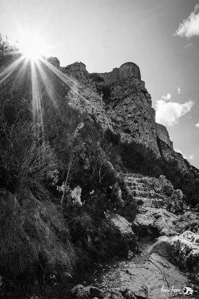 BnW Foto vom Aufstieg zum oberen Teil des oben auf dem gewachsenen Fels gelegenen Château de Peyrepertuse. Die Sonne liegt genau über der Festung und schickt ihre Strahlen zum Tal.
