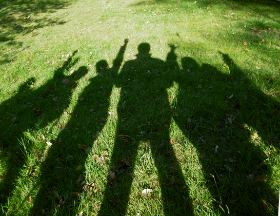 Lange Schatten auf einer grünen Wiese. Zu erkennen sind 5 Personen, eine weitere vesteckt sich im Schatten des Vordermenschys. Die Arme sind ausgestreckt und winken.