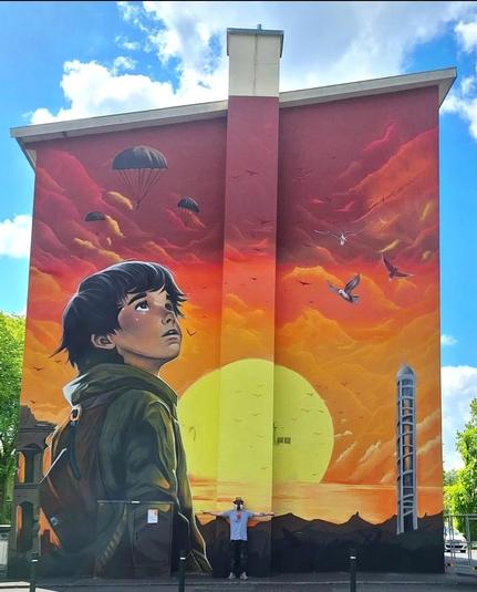 Streetartwall. A monumental 120 m² mural has been sprayed/painted on the outside wall of a multi-storey modern building. The mural is designed in a naive comic painting style and shows a small brown-haired boy in an olive green anorak and backpack in front of a bright sunrise. He is looking up at an orange/red sky in which birds are flying on the right and some parachutists can be seen on the left. A modern long tower completes the mural. (In the photo, the artist is standing in front of his wall.)
Info: On the occasion of the 80th anniversary of the Normandy landings in June 1944, the painting pays tribute to the civilian victims and the resilience of the entire population in an intergenerational form.