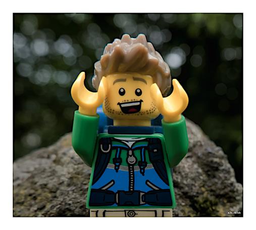 A view of the upper part of the 'hiker' Lego minifigure. The figure has a blue and green torso and a backpack on his back. He has brown spiky hair. His arms are raised in the air and there's a big grin on his face.