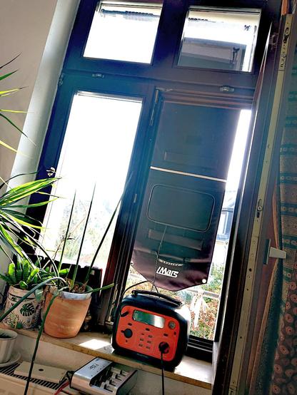 Foto eines Fensters aus dem Innenraum nach außen. Auf dem Fenstebrett (innen) stehen eine orange Powerstation, zwei Grünpflanzen (davon ist eine Sanseveria) . Auf dem Heizkörper unter dem Fensterbrett steht ein Akku-Ladegerät. Einer der beiden Fensterflügel ist geöffnet. Außen hängt (mit der Rückseite zuR BetrachterÏn) ein Solarpanel auf dem iMars steht. Von diesem geht ein Kabel zur orangen Powerstation. In dieser steckt wiederum ein Stromkabel, das möglicherweise zum Akkuladegerät führt. Außen scheint die Sonne. Kastanienbaum und Teile eines anderen Hauses sind erkennbar. Oben im Fensterrahmen ist außen eine Klimmzugstange festgeklemmt, an der das Panel hängt. Unten ist es mit Bändern an der Nicht-Drüberfall-Stange des Fensters festgebunden, so dass es bei Sturm nicht herumflattern kann.