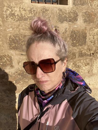 A 50 something trans girl look into the camera. She is wearing large brown sunglasses, her pink hair is in a bun.
She is wearing a pink outdoor jacket and a colourful scarf