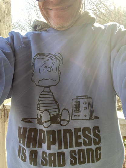 Close up 🆙 f my blue UNIQLO sweatshirt with a picture of Linus crying as he listens to a portable radio. Underneath is the text: “HAPPINESS IS A SAD SONG”