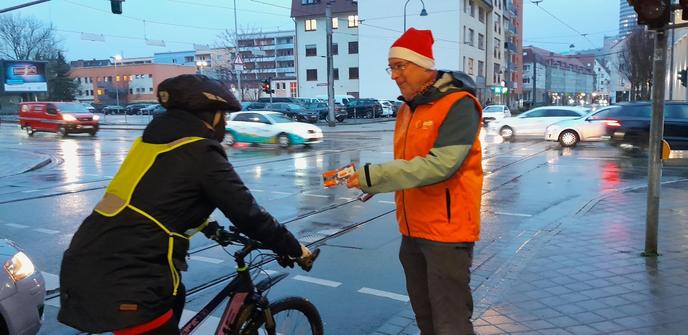 Ein ADFC-Mitglied mit orangefarbener ADFC-Weste und rot-weißer Mütze überreicht einer radfahrenden Person einen Zimtstern. Im Hintergrund ist eine regennasse Kreuzung mit einigen PKW zu sehen.