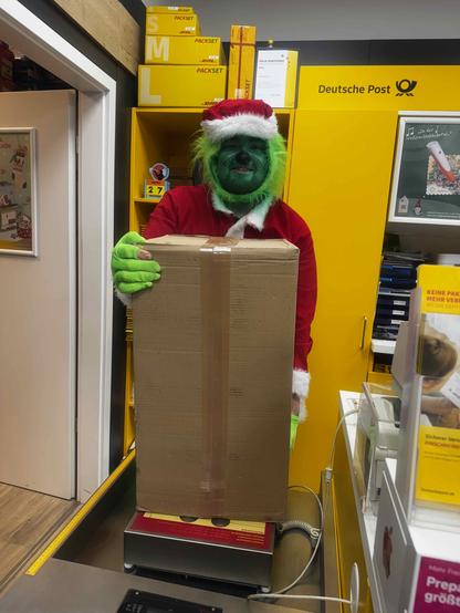Der Grinch im Weihnachtsmannkostüm steht im DHL Shop und hält ein großes braunes Paket in den Händen. 
Und er lächelt dabei!