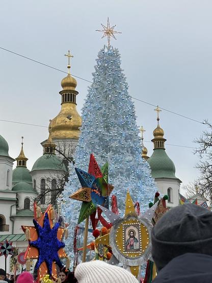 The Ukrainian National Christmas Tree in Sophia Square. It’s about 6 meters tall, white with tiny blue lights and a gold star on top. In the background are the gold and green domes of Sophia Cathedral. The sky is overcast. In the foreground are some of the many homemade stars affixed to the top of long sticks carried by marchers. The stars are about 50 cm across, very colorful, with many points, some decked out in little lights, one bearing an icon of Jesus.