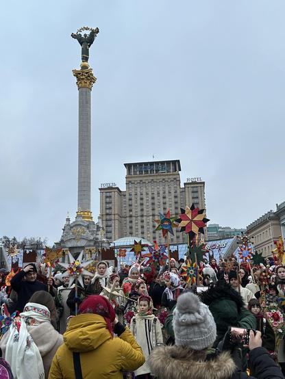 The marchers holding up their stars at the foot of the Independence Monument on the famous Maidan Nezalezhnosti (Independence Square). The monument is 60 meters tall. It’s a column topped by a statue of a woman holding a guelder-rose branch over her head. The sky is overcast.