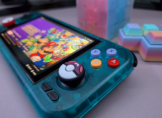 Closeup of the right side of a Retroid Pocket 4 Pro in the translucent Ice Blue colourway. 

The right thumbstick has a purple-white Pokemon Master Ball thumb cap on it, and the ABXY buttons have been swapped out from their stock black background with colour text (red A, yellow B, green Y, blue X) for buttons where the background is colourful. The color scheme is an eclectic mix of red A, yellow B, and light purple X and Y. So basically, US X&Y, and EU A&B.

The screen is showing a colourful still image of a cartoon interpretation of what may or may not be well-known N*ntendo characters.

In the background, a blurry jelly rubiks cube, and a rainbow-coloured Tetris t-piece.

The vibe of the image is colourful, happy, cartoony, and retro-handheld-y.