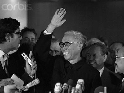 Tho waves his hand while addressing reporters during a press event. He is wearing glasses and a dark outfit, surrounded by various people, including other officials and journalists, who are holding microphones and recording devices.
Black and white photo.