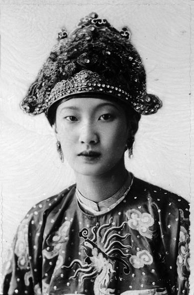 Nam Phuong Empress, wearing a lavish, elaborately decorated Queen headdress with intricate designs and embellishments. Her clothing displays a detailed phoenix pattern in the fabric, which contrasts with the background. The woman’s facial features are prominent, highlighting her serious expression, with natural lighting emphasizing the textures of her attire and accessories. The portrait focuses on her expression and traditional attire.