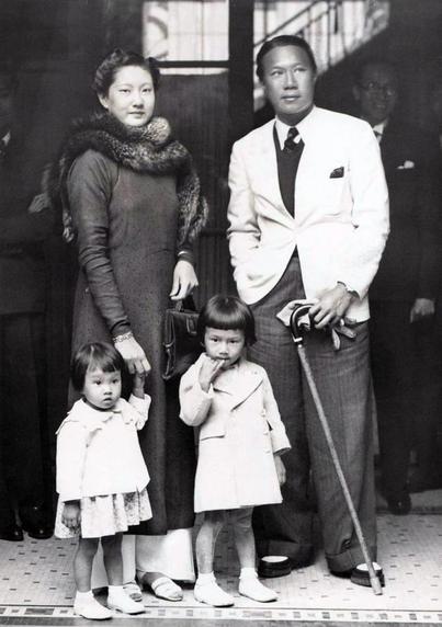 Emperor & Empress with two of their children.
Black and white photo of a family of four standing inside a doorway. The adult couple stands at the center, with the woman wearing a dress and a fur shawl, and the man dressed in a white suit holding a cane. Two young girls, wearing light-colored coats, are positioned in front of them. One girl, to the left, appears to be slightly shy, while the girl on the right poses confidently with her hand raised.