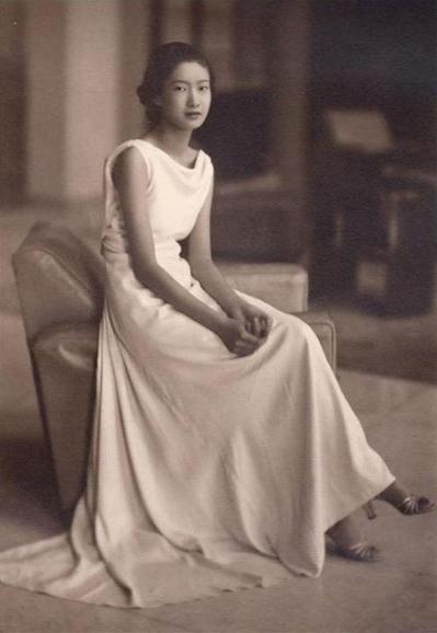 Nam Phuong Empress, in a vintage-style, sleeveless white gown that cascades elegantly over a chair. She is seated in a relaxed pose, with her right hand resting on her left knee and her left hand gently clasped. The photograph has a sepia tone, enhancing its nostalgic feel, with a blurred background showcasing out-of-focus furniture and soft lighting, creating a serene atmosphere.