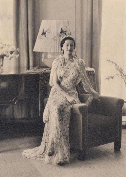 Nam Phuong Empress, seated on side of cushioned armchair, wearing a delicate, patterned dress that falls to the floor. Her hair is styled in an elegant updo, and she gazes softly at the camera. The background consists of softly draped curtains in muted tones, and a lamp with a floral design stands on a dark wooden side table. Subtle plant elements are visible, contributing to a warm, inviting ambiance, with soft lighting that enhances the overall elegance of the scene.