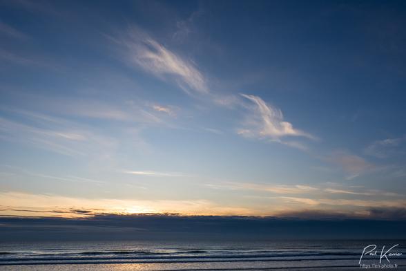 Photo couleur, présentant une fin de journée au-dessus de la mer, captée depuis la plage. De bas en haut, on peut voir le sable et la mer, calme occupant un huitième de la photo, puis une bande nuageuse au-dessus de l'horizon derrière laquelle disparaît le soleil, occupant également un huitième de la photo, puis le reste est occupé par un ciel, essentiellement bleu, avec quelques nuages de très haute altitude, filamenteux, dont trois apparaissent avec des couleurs nacrées peu contrastées.