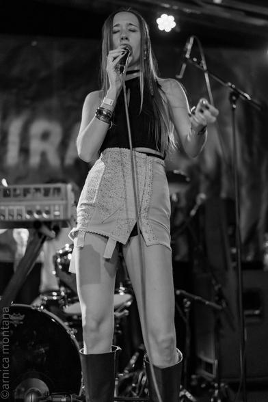 A performer sings into a microphone on stage, wearing a black crop top and a patterned skirt. The image is in black and white, highlighting the stage atmosphere, with musical instruments visible in the background.
