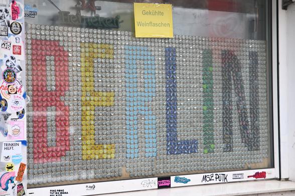 A shop window display made of bottle caps arranged to spell "BERLIN" in colorful letters. The background consists of silver bottle caps, while the letters are formed using red, yellow, blue, green, and black caps. A yellow sign above the display reads "Gekühlte Weinflaschen" (Chilled Wine Bottles). The window is decorated with various stickers and graffiti.