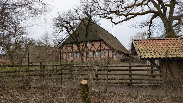 An old-fashioned farm house with bricks, timber frame and a thatched roof; a fence; bare trees.