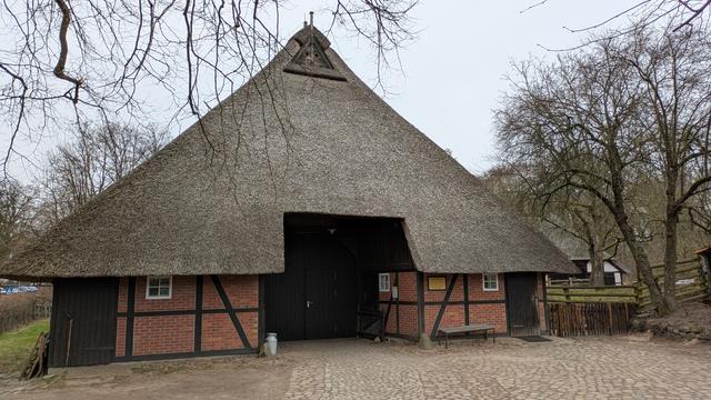 A timber frame barn with a large thatched roof. Bare trees.
