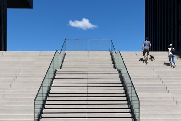 Look-up view of concrete minimalist stairs between two dark modern buildings under a blue sky.  Two greenish transparent glass ramps in the middle of the stairs attract the viewers' attention to a tiny white cloud in the sky. A couple with a little dog is about to arrive at the top of the stairs on the right side. The shadows of their tiny silhouettes also lead in the cloud direction.