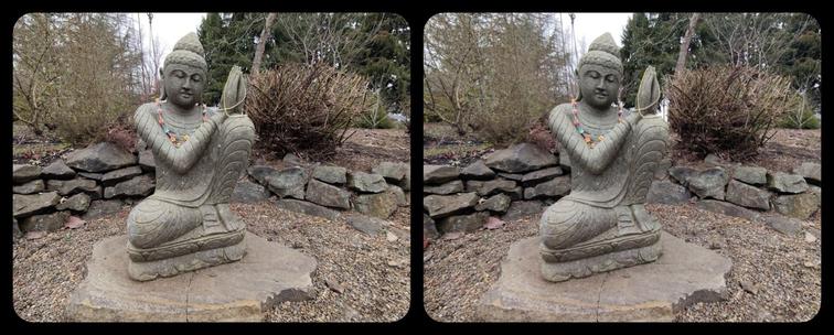 Stereoscopic, parallel view. Outdoors. A seated Buddha sculpture, made of stone. Praying hands held on left side, left knee up. Decorated with a colorful necklace and some gold-colored beads draped around the hands. There is a low, semicircular wall of irregular stones behind.