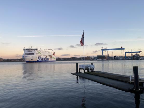 Eine Stena Fähre bei der Ausfahrt aus der Kieler Förde. Die Fähre hat den Bug herumgedrückt. Blick von der Kiellinie, im Vordergrund einer der Ruderstege, im Hintergrund die Werftkräne am Ostufer. Die See ist glatt, der Himmel am höchsten Punkt bläulich, zum Horizont hin rötlicher mit einigen Wolkenstreifen.