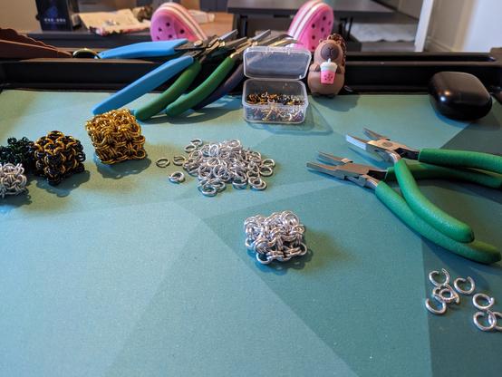 My work station, with a collection of chainmaille fidget cubes of various designs to left, my pliers to the right, and pile of silver rings in the middle with a partially constructed silver fidget cube in front. In the background, my plastic capybara, Captain Barry, keeps watch.