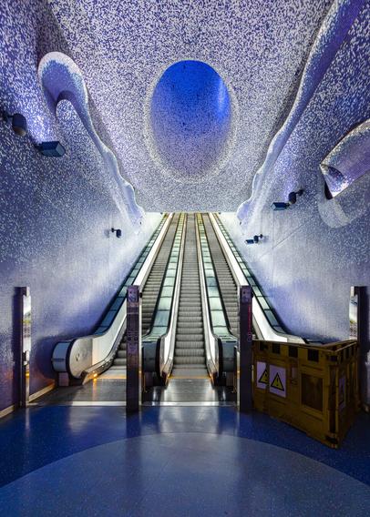 Vertical look-up view of a set of three very long escalators beneath a gorgeous ceiling mosaic painted in a psychedelic swirl of violet-blue. The Walls are also covered with thousands of blue mosaic tiles and give the feeling of being immersed in the sea. The escalators are surrounded by a huge cone of light that extends from the top to bottom of the station and bathes the space in a surreal blue light at this very moment (the color of this light cone is overwise changing constantly from purple to blue and green every fews seconds). The floor is also blue. The escalators where free of people when the photographer took the shot. The escalator on the right was unfortunately out of order on that day and access to it was prevented with a mobile yellow plastic fence.