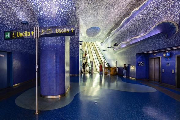 This third photo was taken from even further behind and includes an impressive pillar also covered in mosaic and looking similar to a water fountain on the left of the image in the foreground. There is also a metal pole with entry and exits signs in italian.  This time too, a few people are visible in the escalators area.