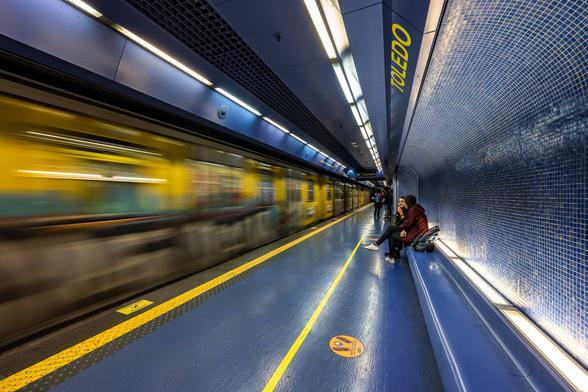 View on one platform of the The Toledo metro station featuring a yellow metro train covered in graffiti entering a deep blue station. The walls on the right form the shape of a curve reminding of a wave on the top and are covered in tiny dark blue mosaic. Below  this "wave" two young ladies are waiting for the train to stop seating on a very long dark blue plastic bench.