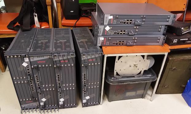 Picture shows seven rack mount devices.
Three 2u Avaya G430's are piled on a chair with a helixal 1800Mhz antenna under it and a SmartStore box with a threephase plug in it.
Four 4U or 5U Avaya G450's stand on their side next to it.