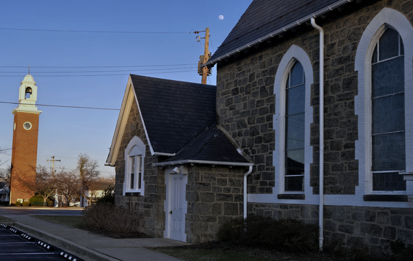 Small distant near-full moon at upper center over a beautiful 19th-century stone chapel. To its left and in the background is a stand-alone red brick belltower several stories tall (with some Moiré pattern aberration). Evening light with the sky a dimming blue, no clouds. We see the left back end of the chapel, showing a smaller window and white wooden door. Toward the right of the frame are two large stained glass windows in pointed arch frames. Mood is quiet, admiring the antique stonework and evening coming on. 

Historic St. Mark's Catholic Church, 2025 Mar 11 06:30 pm EDT. Color photo by @KronoMoon

www.KronoMoon.org 🏳️‍🌈 ☮️ 
#KronoMoon #MoonOverMastodon #KronoWatch