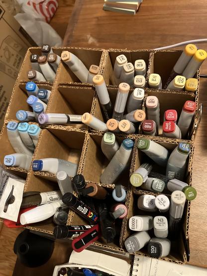 View from above of a homemade upcycled cardboard storage container of Copic ink refill tubes, organized into separate compartments by color family, along with some Posca markers and Sharpie paint markers.