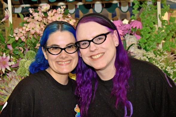 The image features two women standing side-by-side, facing the camera, with their heads touching. The woman on the left has blue hair styled in a ponytail, while the woman on the right has long purple hair that falls down her back. Both individuals are wearing black shirts and have dark-framed glasses.

In the background, there is a blurred floral arrangement featuring pink flowers, green leaves, and possibly white blooms. The overall atmosphere suggests a casual, relaxed setting, likely taken in a home or garden environment.