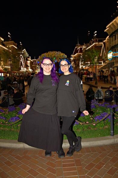 The image features two women standing together in front of a lush greenery backdrop, posing for the camera. The woman on the left has long purple hair and is dressed in a black shirt with three-quarter-length sleeves, paired with a long black skirt that falls just above her ankles. Her counterpart on the right sports short blue hair and wears a black shirt with a white logo on the front, complemented by black pants.

The duo stands on a brick pathway, surrounded by vibrant purple flowers and lush greenery behind them. In the background, several buildings are visible, including one adorned with lights along its roofline. The dark sky above suggests that the photo was taken at night, adding to the overall ambiance of the scene.