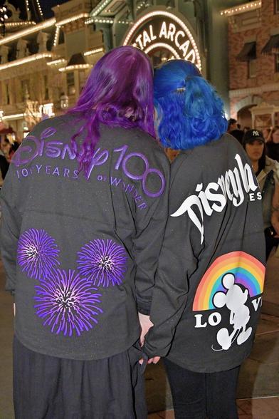 The image shows two people standing together, facing away from the camera, with their backs to the viewer. The person on the left has purple hair and is wearing a black long-sleeved shirt with a large purple flower design on the back. The person on the right has blue hair and is wearing a black long-sleeved shirt with white text that reads "Disney 100 Years of Wonder" across the shoulders, along with a rainbow-colored Mickey Mouse logo on the lower back.

In the background, there are several buildings visible, including what appears to be an arcade or game center. The overall atmosphere suggests that the two individuals are likely at Disneyland or another Disney-themed park, given their attire and the presence of Disney branding in the background.