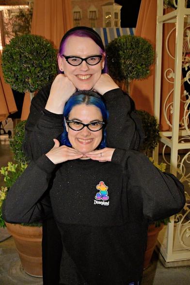 The image features two women posing together, each wearing black sweaters adorned with colorful logos on the left side of their chests. The woman in front has blue hair styled into a bun at the back of her head, while the woman behind her sports purple hair and rests her chin on the other's shoulder.

In the background, a potted plant sits atop a table or shelf, accompanied by an orange curtain hanging from a white metal frame to the right. The overall atmosphere suggests that this image was captured in a public setting, possibly at a convention or event where attendees were encouraged to take photos with friends and family.