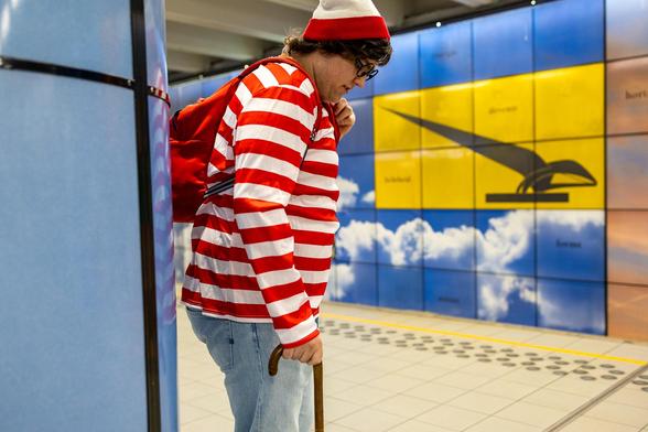 A man dressed in a red and white striped T-shirt and jeans and wearing a red and white striped woollen hat and a red rucksack, just like the Wally charachter in the popular series of children’s puzzle books by British illustrator Martin Handford. is standing behind a blue pillar on a metro platform in brussels. He is looking at his shoes as if he had been caught. Opposing him, on a wall, the drawing of a black bird on a yellow backround is pointing his tail at the man's nose. Wally is also known as Waldo in the US, Charlie in France or Walter in Germany.