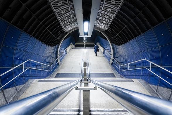 Look-down shot with a wide-angle lens of stairs going down to underground platforms through a dark blue, black and grey tube with a Sci-Fi look. The silhouette of a man with a dark blue suit appears on the right at the bottom of the first set of stairs.