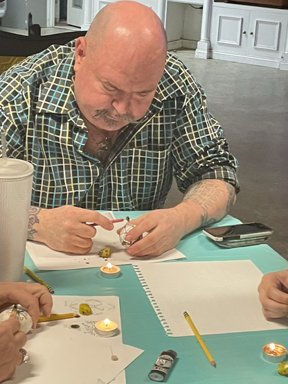 A man writing pysanky eggs. With a blue table cloth with eggs, paper, pencils, candle and kitskas
