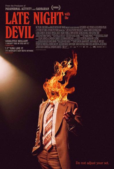 Poster for the 2023 Cameron Cairnes and Colin Cairnes motion picture “Late Night with the Devil.” The image shows a typical late-night talk show host in a spotlight, wearing a suit and tie, gesturing towards the unseen audience; but his head has been entirely replaced by flames.