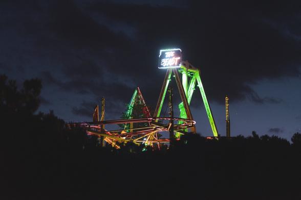 The image shows a nighttime scene featuring a brightly lit amusement park ride or roller coaster called "The Beast." The ride is illuminated with colorful lights, and a neon sign with the name is visible on top. The sky is dark with some clouds, creating a dramatic backdrop for the ride.