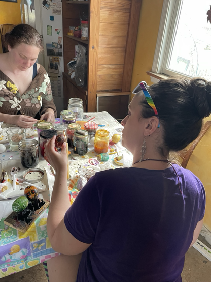 Two women writing pysanky.  Colorful eggs and dyes are on a table.