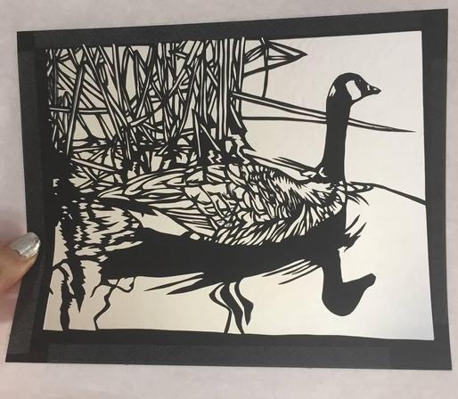 Black paper painstakingly cut so that a design of a Canada goose swimming past reeds shows.