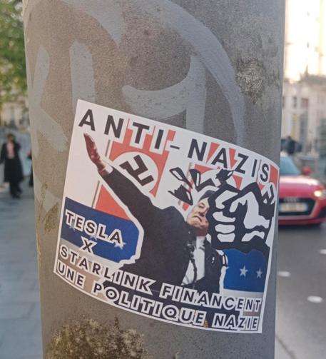 Un autocollant sur un poteau qui montre elon musk faisant le salut nazi avec un coup de poing qui le frappe a la figure. Est noté "anti- nazis tesla x Starlink financent une politique nazie"