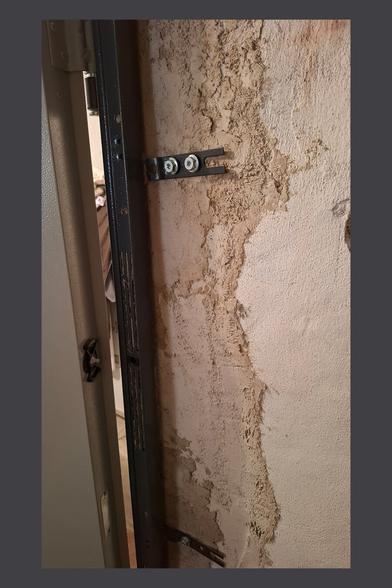 This image shows a close-up of the door frame and its mounting structure. The door is slightly open, revealing the metal frame and the wall behind it. The wall has a rough plastered surface with an uneven texture, indicating prior renovation work.
Two black metal brackets are secured to the wall with screws, helping to anchor the door frame. The brackets have an open slot design, possibly to allow for adjustment before final tightening. The frame itself appears to be a sturdy metal construction with ventilation or drainage slots.
This view gives insight into how the door was fixed into the wall, showing both the structural integration and the unfinished nature of the surrounding plasterwork.
