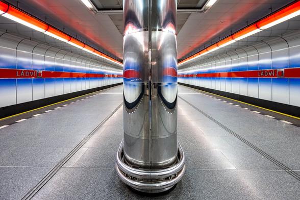Wide-angle symmetrical shot of a metro platform with white, blue and red stripes at the Ladvi metro station in Prague. Those stripes are reflected in a big round metal pillar with a mirror effect in the foreground of the image creating two sorts of googly eyes.