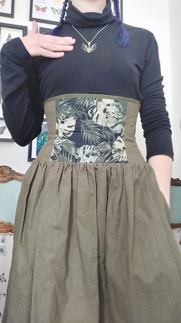 Plan rapproché sur ma tenue du jour. 
Je porte un haut noir à manches longues avec un col montant, et une jupe corsetée que j'ai réalisée.
La partie corsetée commence sous la poitrine et va jusqu'au dessus des hanches. Elle est composée d'un panneau devant en coton noir avec des motifs de feuilles tropicales dans un camaïeu de vert. Les panneaux sur les côtés sont en lin vert kaki, comme le reste de la jupe. 
On ne le voit pas, mais elle est lacée dans le dos, ce qui permet de l'ajuster à sa morphologie. 

L'une de mes mains est posée sur mon buste pour mettre en avant le pendentif que je porte. Il représente une petite fée aux ailes vertes et noires, réalisée par l'atelier le bois des Fées (dont le compte est mentionné dans la publication). 
Mon autre main est cachée dans la poche de ma jupe, car oui, elle a évidemment des poches !

Le fond de la photo est un mur blanc avec des cadres divers.