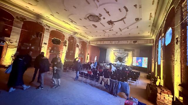 An interior shot of a spacious, partially renovated room with exposed brick walls and decorative ceiling details. Several people mingle in the foreground, surrounded by rows of chairs. A screen displays colorful visuals at the far end, while artistic lighting highlights the room’s features