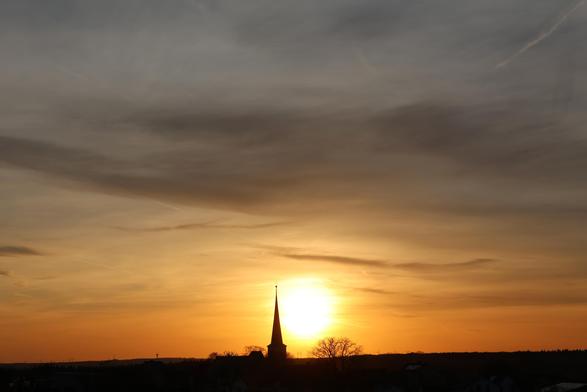 Sonnenuntergang kein blauer Himmel, so denn ein dunkelgrauer in der oberen Hälfte des Fotos. Die weisse Sonne umrahmt von unterschiedlichen Orangetönen. Die Silhouette eines Kirchturmes und eines großen Baumes.
Matzework
