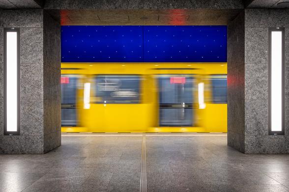 View from the opposite platform of a yellow metro train passing between 2 grey pillars at Museumsinsel U-Bahn Station in Berlin under a painted dark blue starry ceiling.
